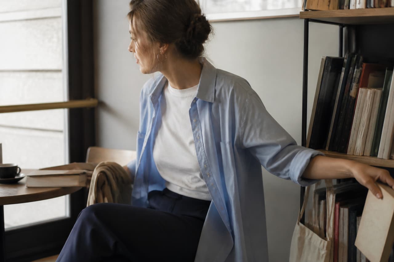 Mujer con camisa azul clara abierta sobre camiseta blanca en una biblioteca.